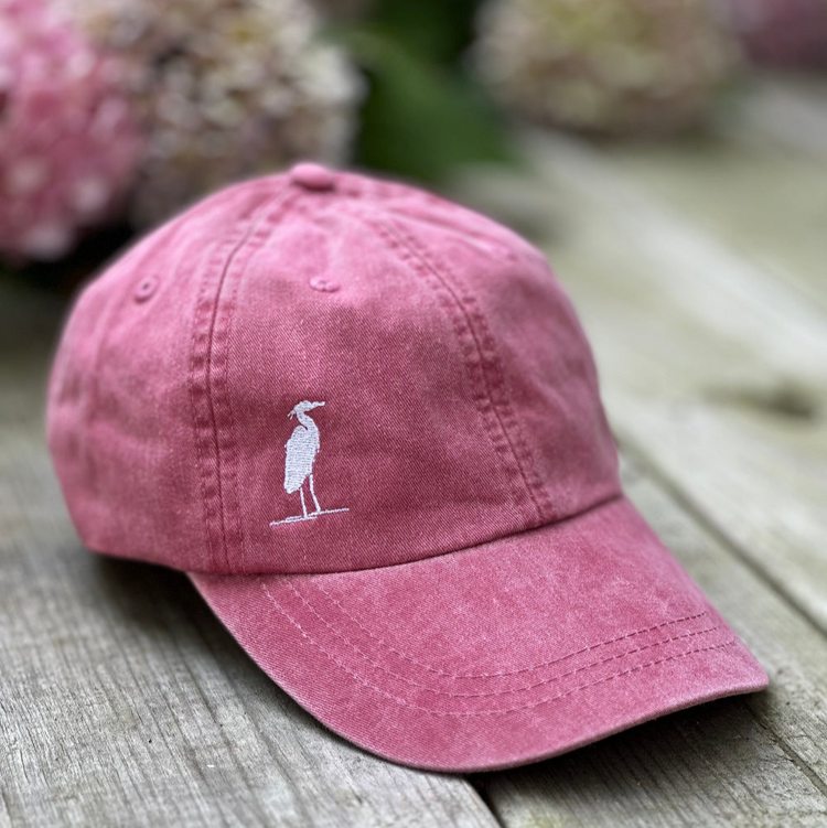 Nantucket Red cap with a White Heron logo on a wooden surface with blurred flowers in the background